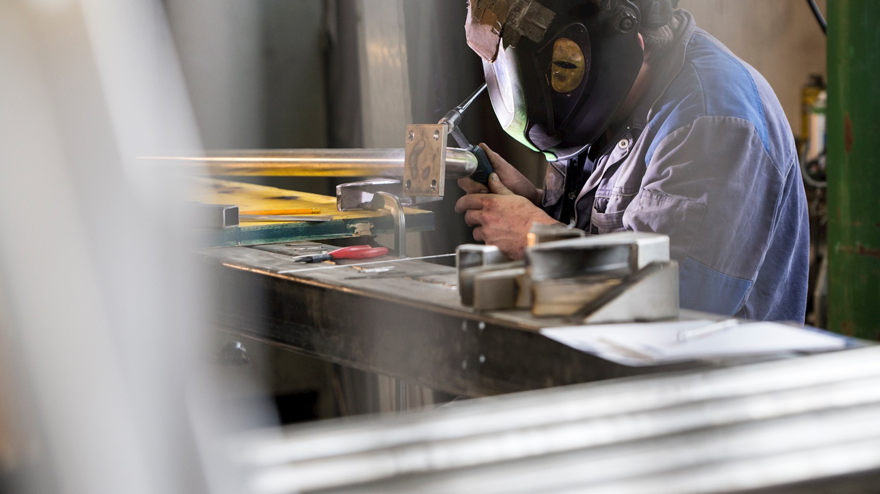 Mann mit Schweißschirm bearbeitet ein Metallteil auf einer Werkbank