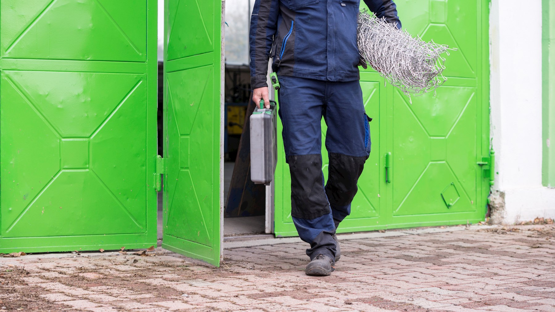 Mann in blauer Arbeitskleidung verlässt die Leo Werkstatt durch eine hellgrüne Tür. Unter seinem linken Arm trägt er eine Rolle Zaundraht. In seiner rechten Hand einen Werkzeugkoffer.