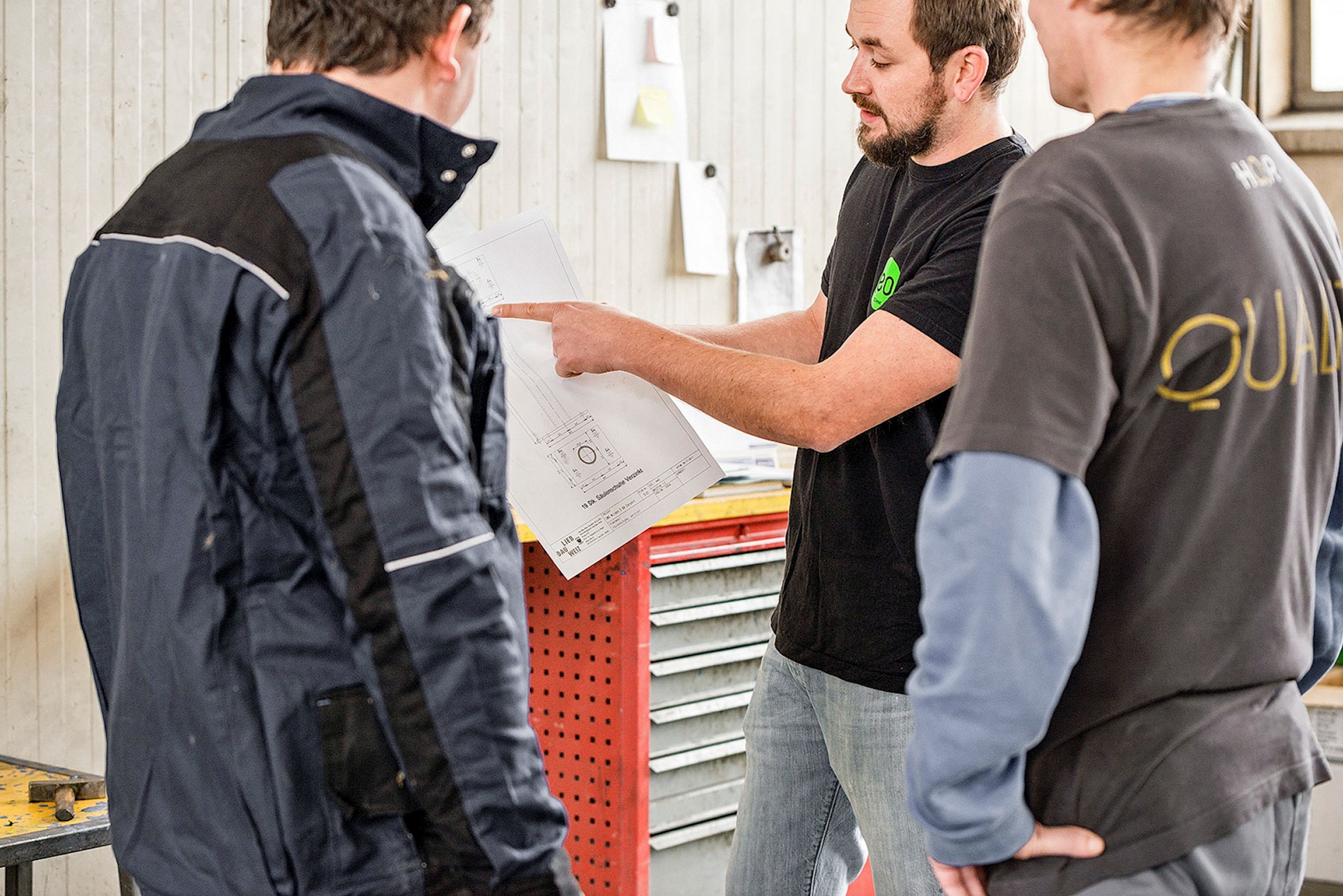 3 men are wearing work clothes and looking at a plan. One man points at the plan and explains something.