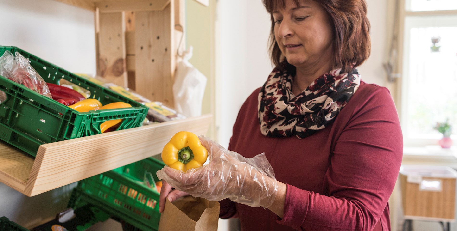 You can see an employee in the social supermarket in Fürstenfeld. She is wearing a plastic glove and putting a yellow pepper into a paper bag.