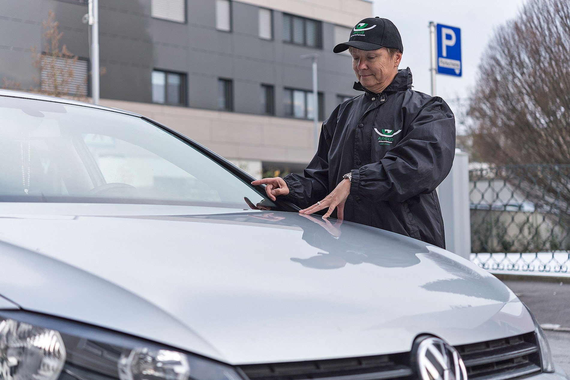 Eine Frau trägt eine schwarze Regenjacke und eine schwarze Kappe mit dem ARGUS-Logo. Sie schaut durch die Windschutz-Scheibe in ein parkendes Auto.