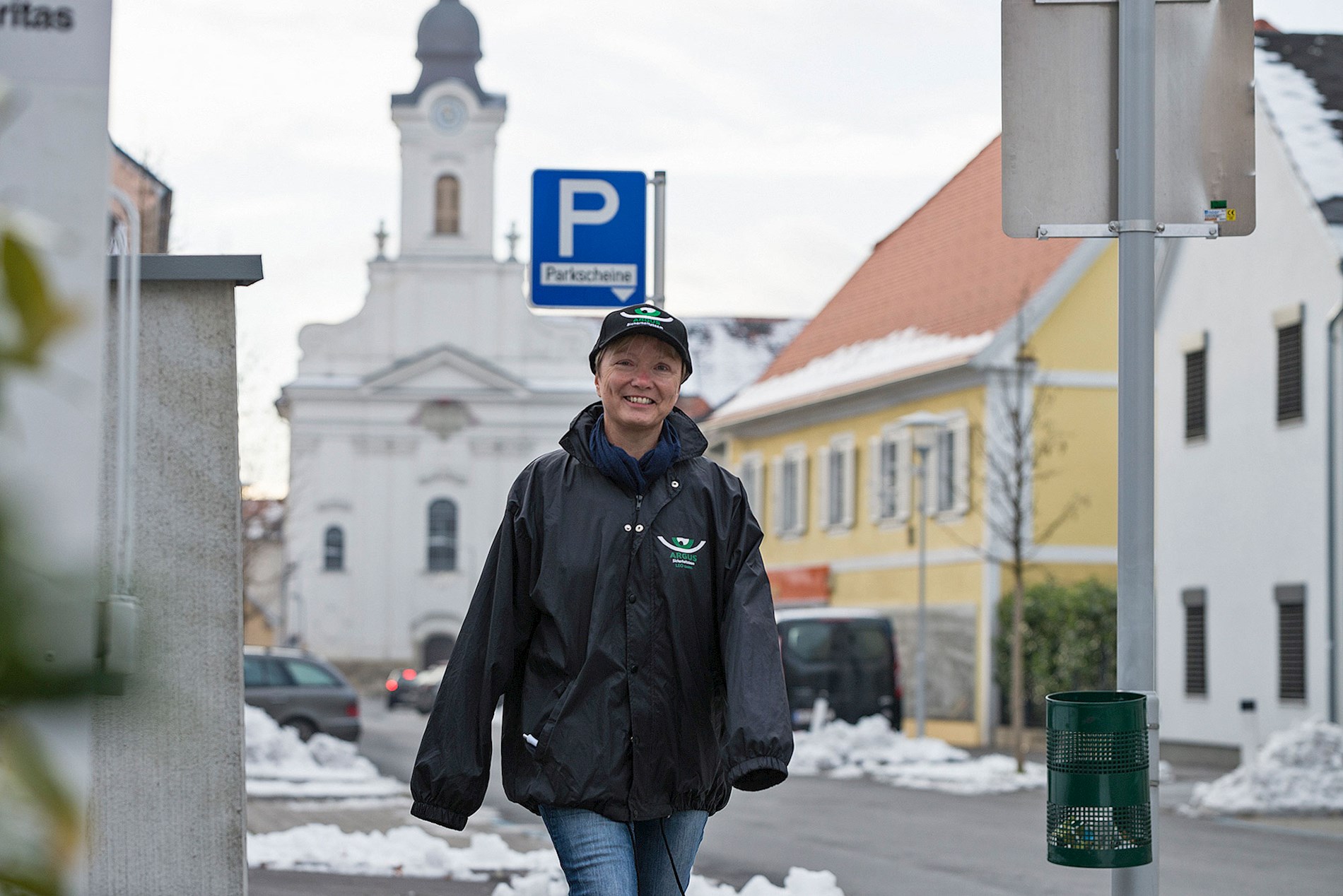 A woman is wearing a black rain jacket and a black hat with the logo of ARGUS. She is smiling and walking down a street towards us.