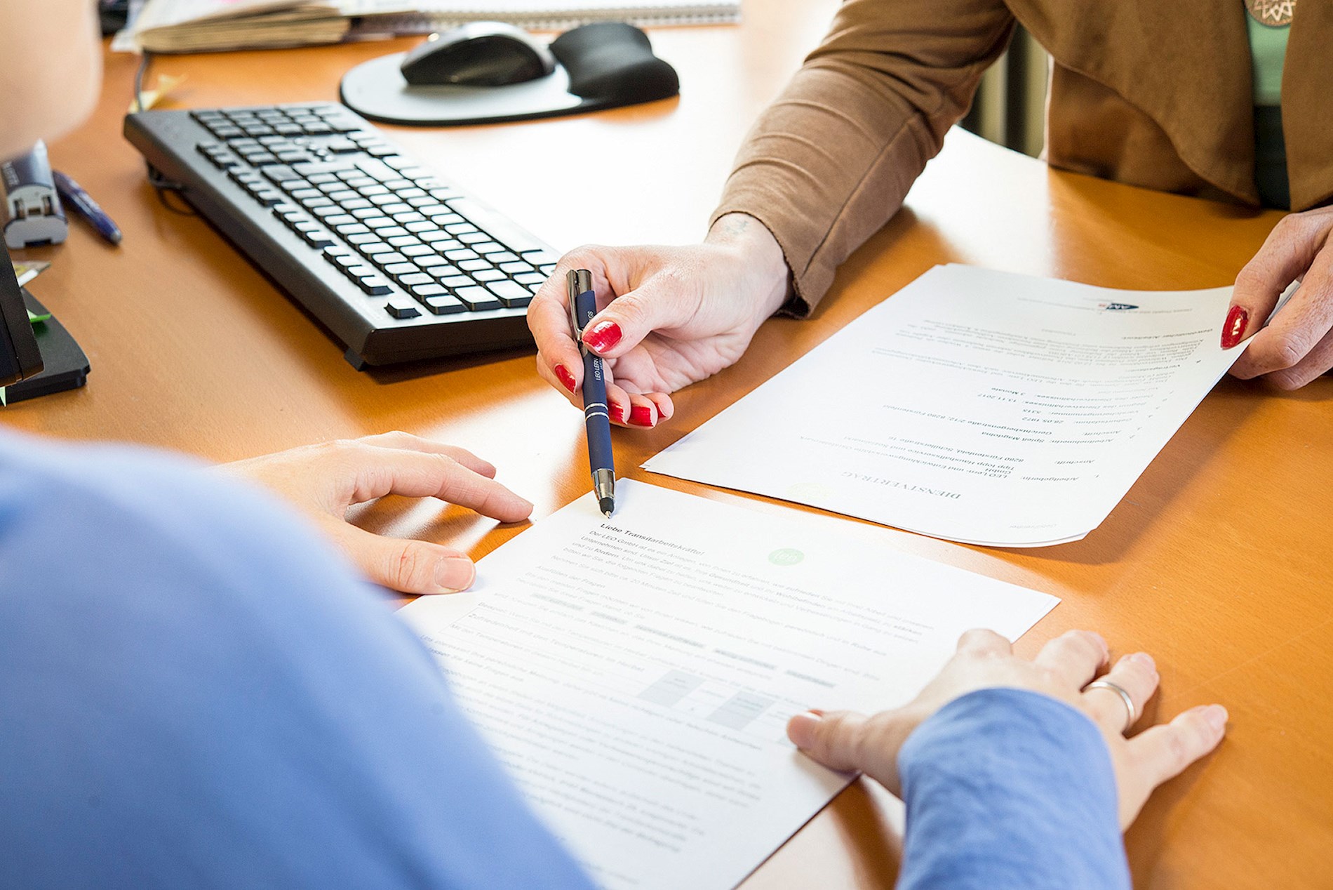 You can see the hands of 2 people at a desk. On the desk, there are printed pages. One person points to a page with a pen.