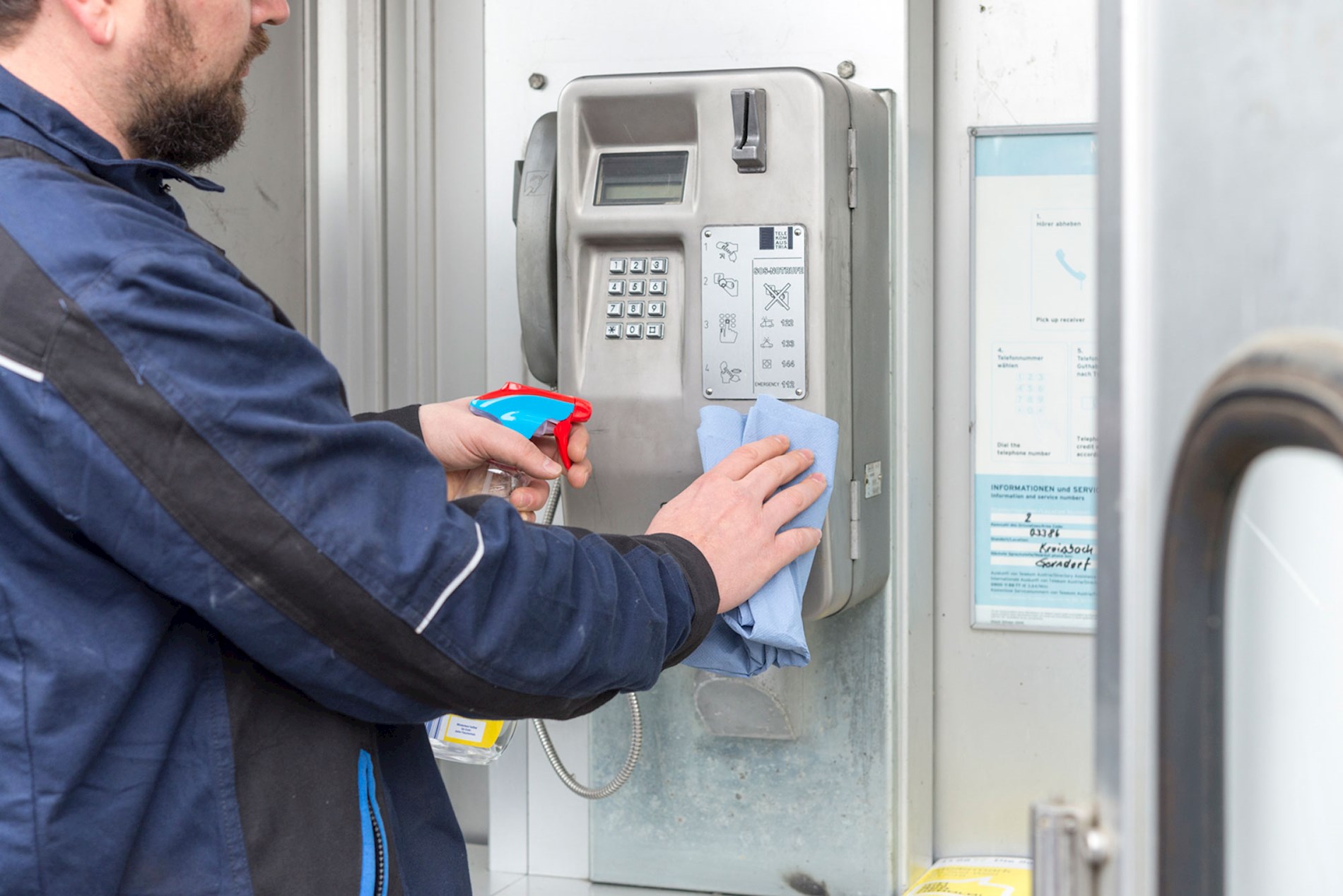 Eine Person putzt das Telefon in einer Telefonzelle. Sie verwendet Putzmittel in einer Sprühflasche und einen hellblauen Lappen.